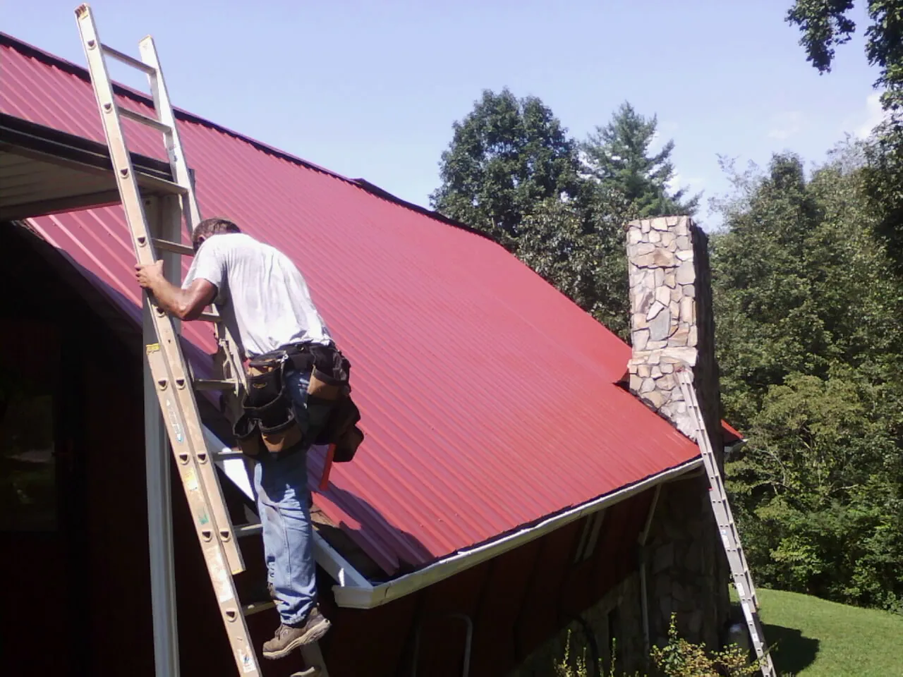 Roofer on a ladder beside a red metal roof and stone chimney — Soffit & Fascia Repair by Roofing Brothers in Woolwich